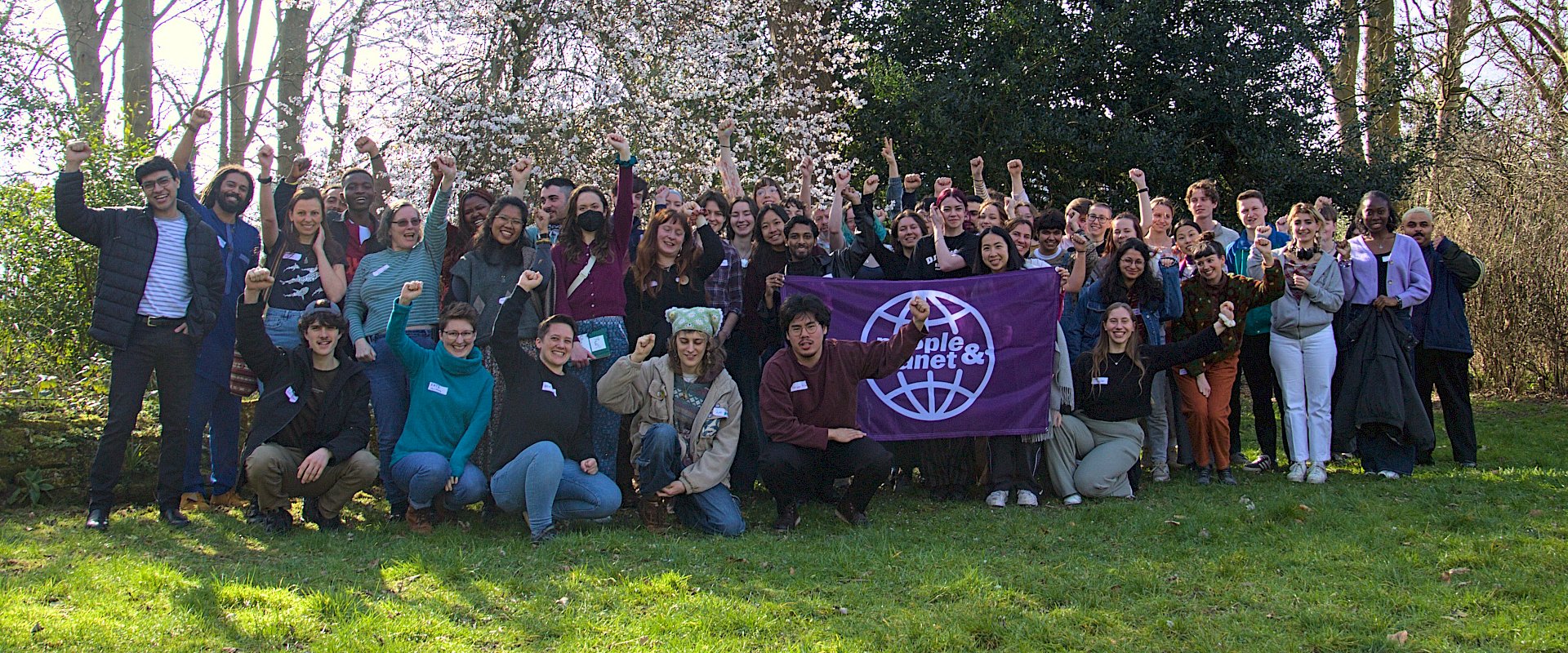 Photo of large group of students holding up a People & Planet banner with fists in the air amongst trees